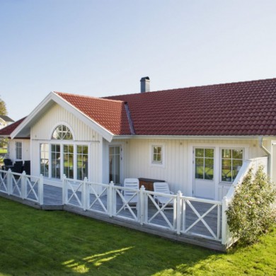 Exteriör av ett vitmålat enplanshus med rött tegeltak. Huset har en bred veranda med vita räcken och flera glasdörrar som leder ut till trädgården. En gräsmatta omger huset, och några träd och buskar syns i bakgrunden. Det är en solig dag med klarblå himmel, vilket ger huset en ljus och inbjudande framtoning.