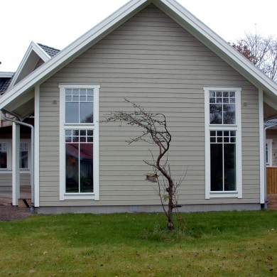 Framsida av ett hus med en spetsig taklinje, målat i beige med vita fönsterkarmar. Två stora fönster, ett på vardera sida av fasaden, ger utsikt över interiören. Framför huset växer ett litet, krokigt träd med få löv. Gräsmattan framför huset är grön, och en röd byggnad syns delvis i bakgrunden.