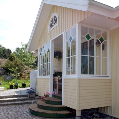 En solig veranda på ett hus med gul träpanel och vita detaljer. Verandan har stora fönster med spröjsade rutor och färgade glasdetaljer upptill. En halvcirkelformad fönsteröverdel pryder verandans gavel. Trappsteg med grön matta leder upp till verandan, och blomkrukor med färgglada blommor står vid ingången. I bakgrunden syns en stenlagd gång och frodig grönska, vilket skapar en inbjudande och somrig atmosfär.