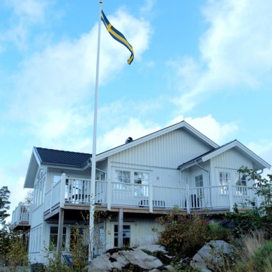 Exteriör av ett tvåvåningshus med vit träpanel, beläget på en sluttning. Huset har flera balkonger med vita räcken och stora fönster som släpper in mycket ljus. En svensk flagga vajar högt på en flaggstång framför huset. Omgivningen består av klippor och växtlighet, och himlen är blå med några vita moln, vilket ger en naturnära och svensk atmosfär.