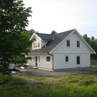 Exteriör av ett vitmålat tvåvåningshus med svart tak, beläget på en rymlig tomt omgiven av grönska. Huset har flera fönster och en dörr som leder ut till en grusad gång. En stor trädgård omger huset, med gräsmattor och blommande växter. En del av tomten är fortfarande under arbete med byggmaterial synligt nära huset. Ett stort träd skymtar i förgrunden och ger skugga till delar av gården.