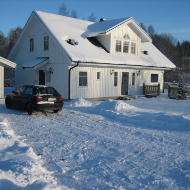 Exteriör av ett vitmålat tvåvåningshus med svart tak, täckt av snö. En snötäckt uppfart leder fram till huset, där en svart bil är parkerad. Huset har en balkong ovanför entrén och flera fönster som är delvis täckta av snö. Snön täcker marken runt huset, och träd i bakgrunden är också täckta av snö, vilket ger en vinterlig och fridfull atmosfär. Himlen är klarblå, och solen skiner, vilket skapar långa skuggor på snön.